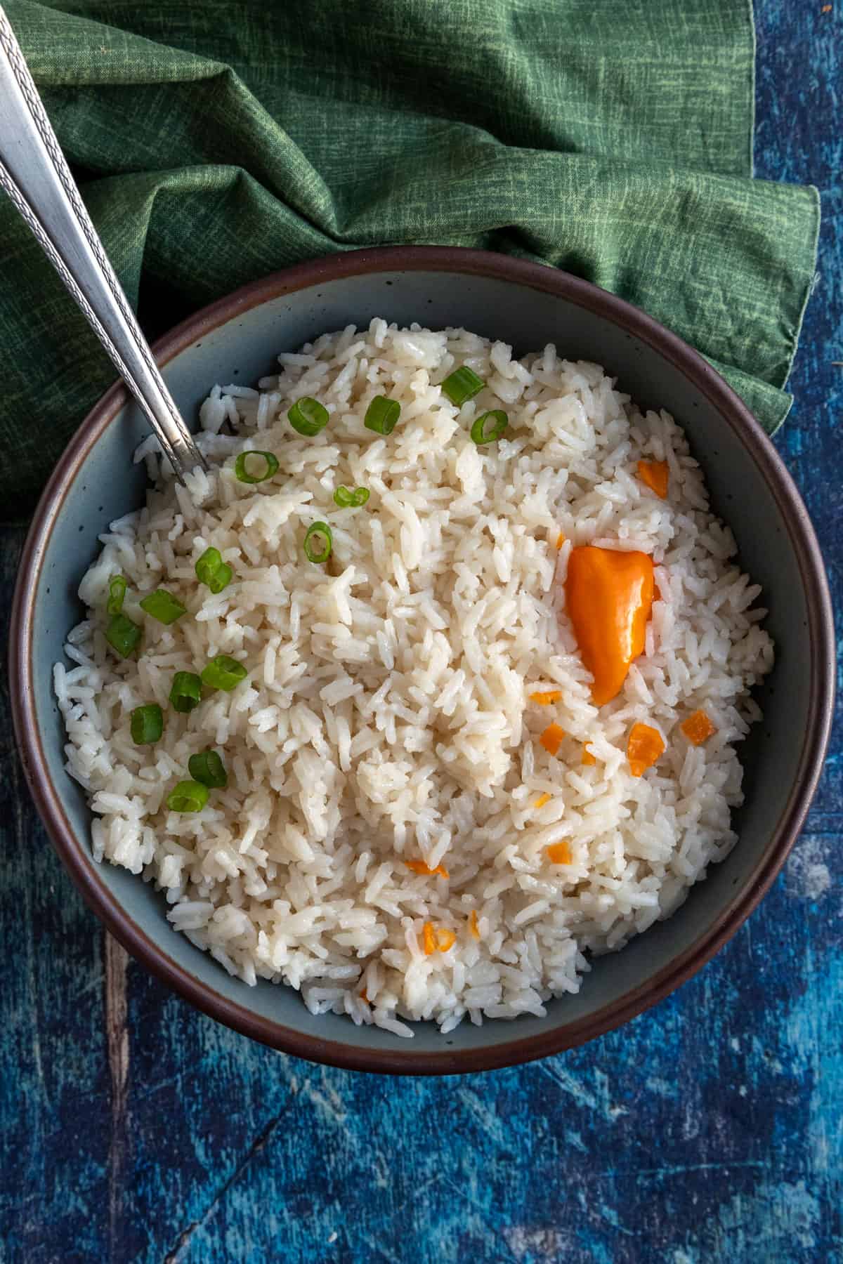 A bowl of coconut rice garnished with chopped green onions and orange habanero peppers sits on a blue surface, with a green cloth and a spoon resting in the bowl.