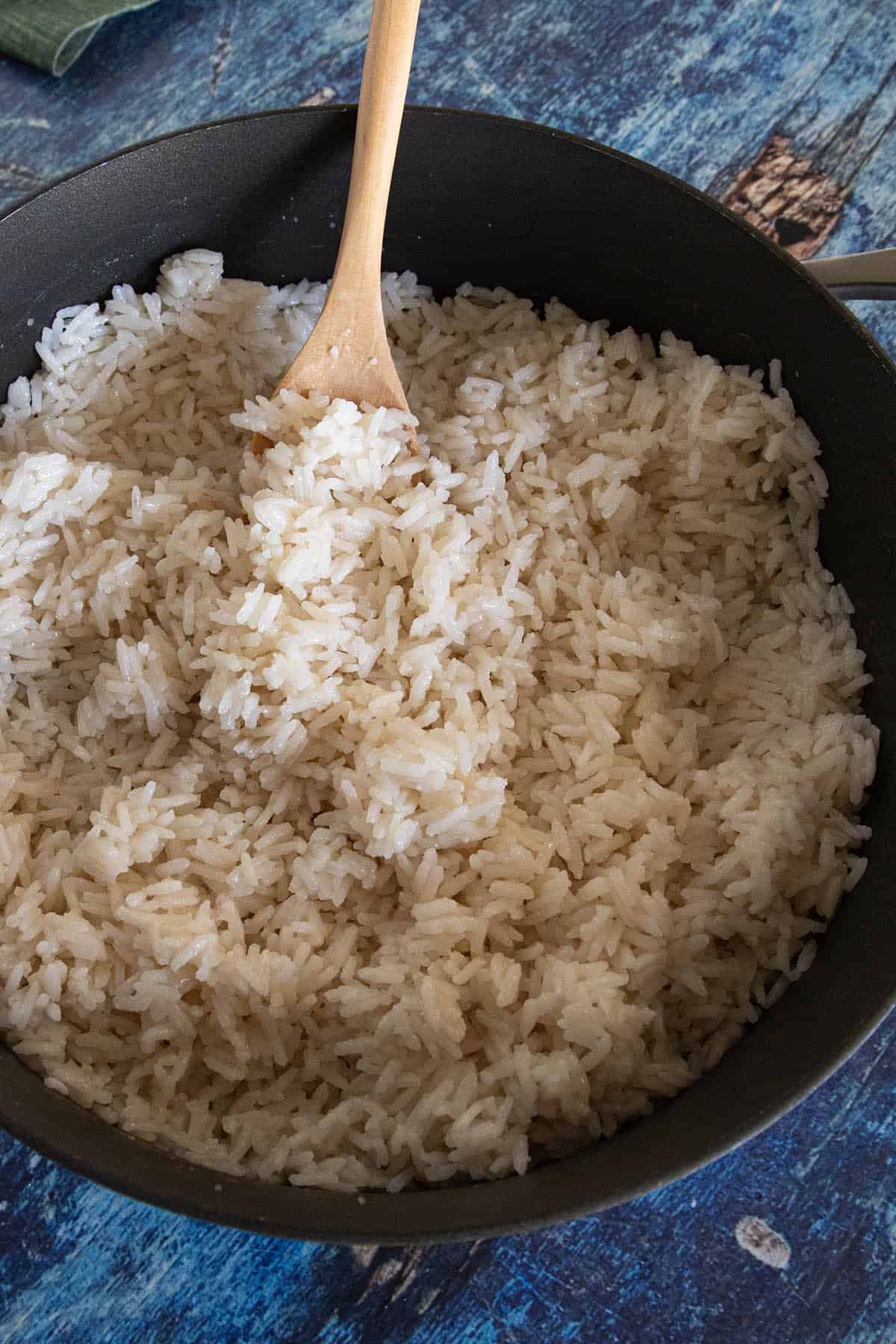 A pan filled with freshly cooked coconut rice being stirred by a wooden spoon, sitting on a blue textured surface.