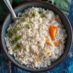 A bowl of creamy coconut rice garnished with chopped green onions and mixed with orange habanero pieces. A spoon rests in the bowl, which sits on a blue wooden surface next to a green napkin.
