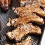 A close-up of juicy, oven baked ribs arranged on a metal baking tray. The ribs are golden brown with a glazed surface, showing tender meat and bones.