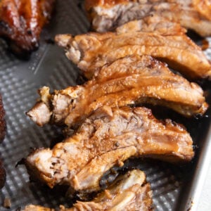 A close-up of juicy, oven baked ribs arranged on a metal baking tray. The ribs are golden brown with a glazed surface, showing tender meat and bones.