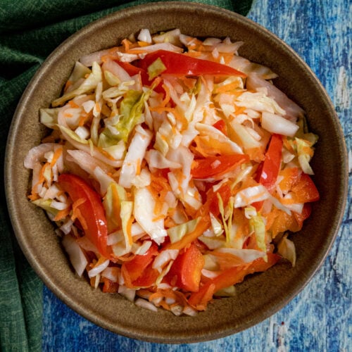 A small bowl of fresh Pikliz (Spicy Haitian Pickled Slaw with shredded cabbage and chilies) is placed on a blue surface next to a green textured cloth.