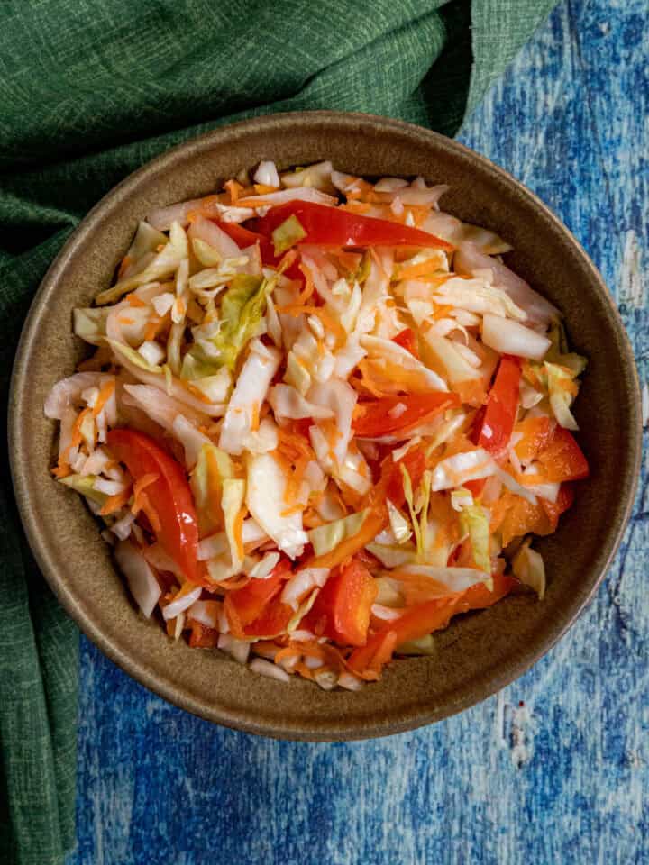 A small bowl of fresh Pikliz (Spicy Haitian Pickled Slaw with shredded cabbage and chilies) is placed on a blue surface next to a green textured cloth.
