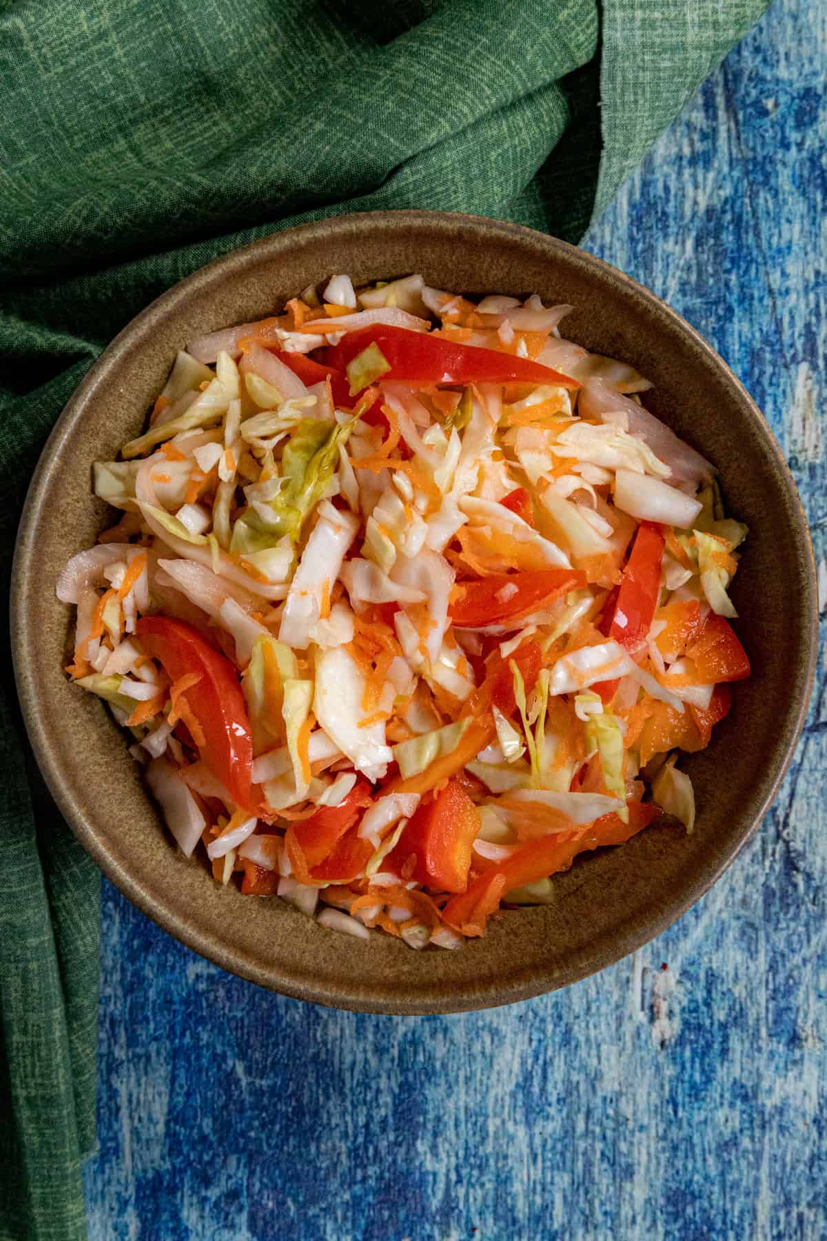 A small bowl of fresh Pikliz (Spicy Haitian Pickled Slaw with shredded cabbage and chilies) is placed on a blue surface next to a green textured cloth.