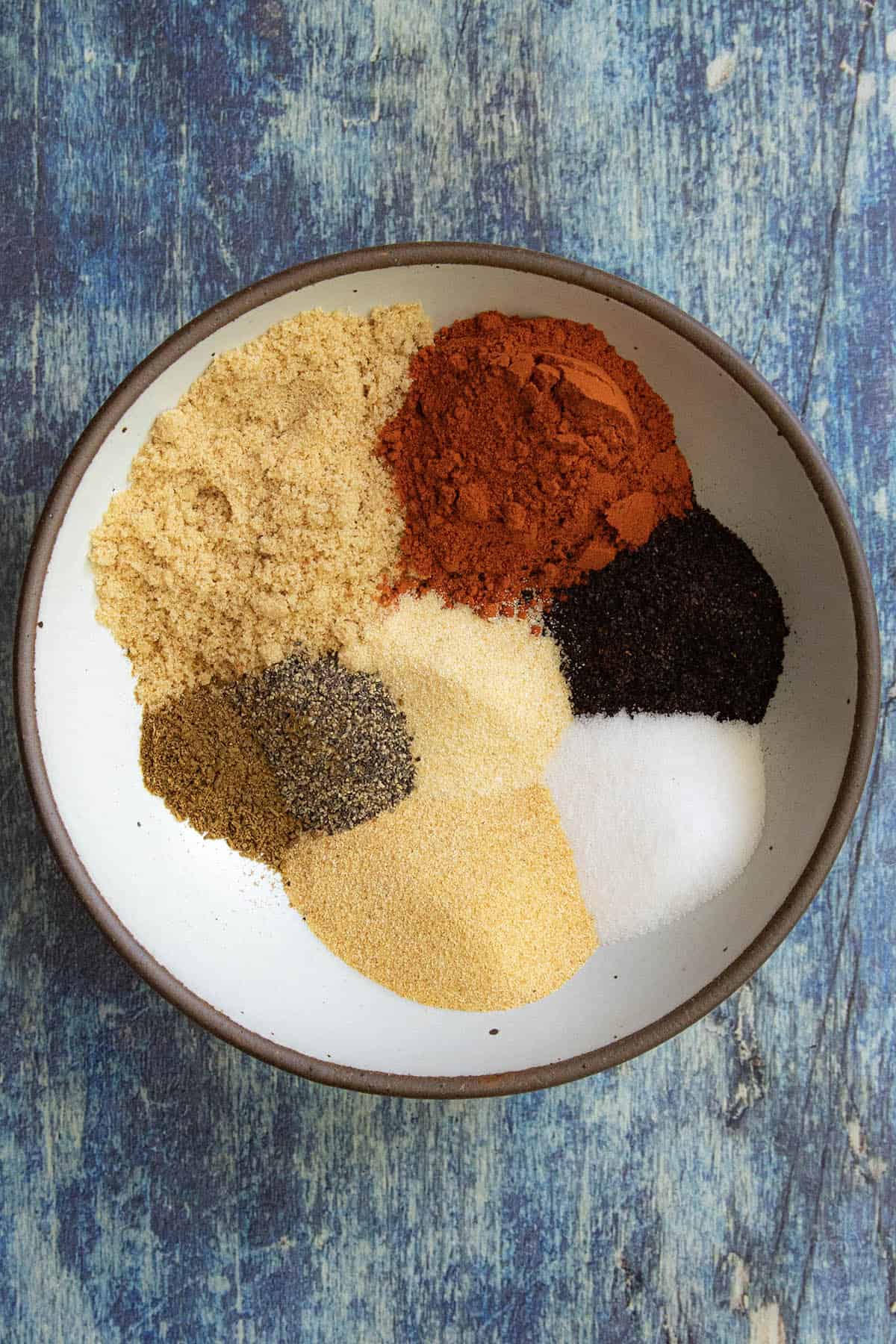 A bowl containing neat piles of various ground spices, perfect for a flavorful rib rub, including red, yellow, black, and white powders, sits on a blue textured surface.