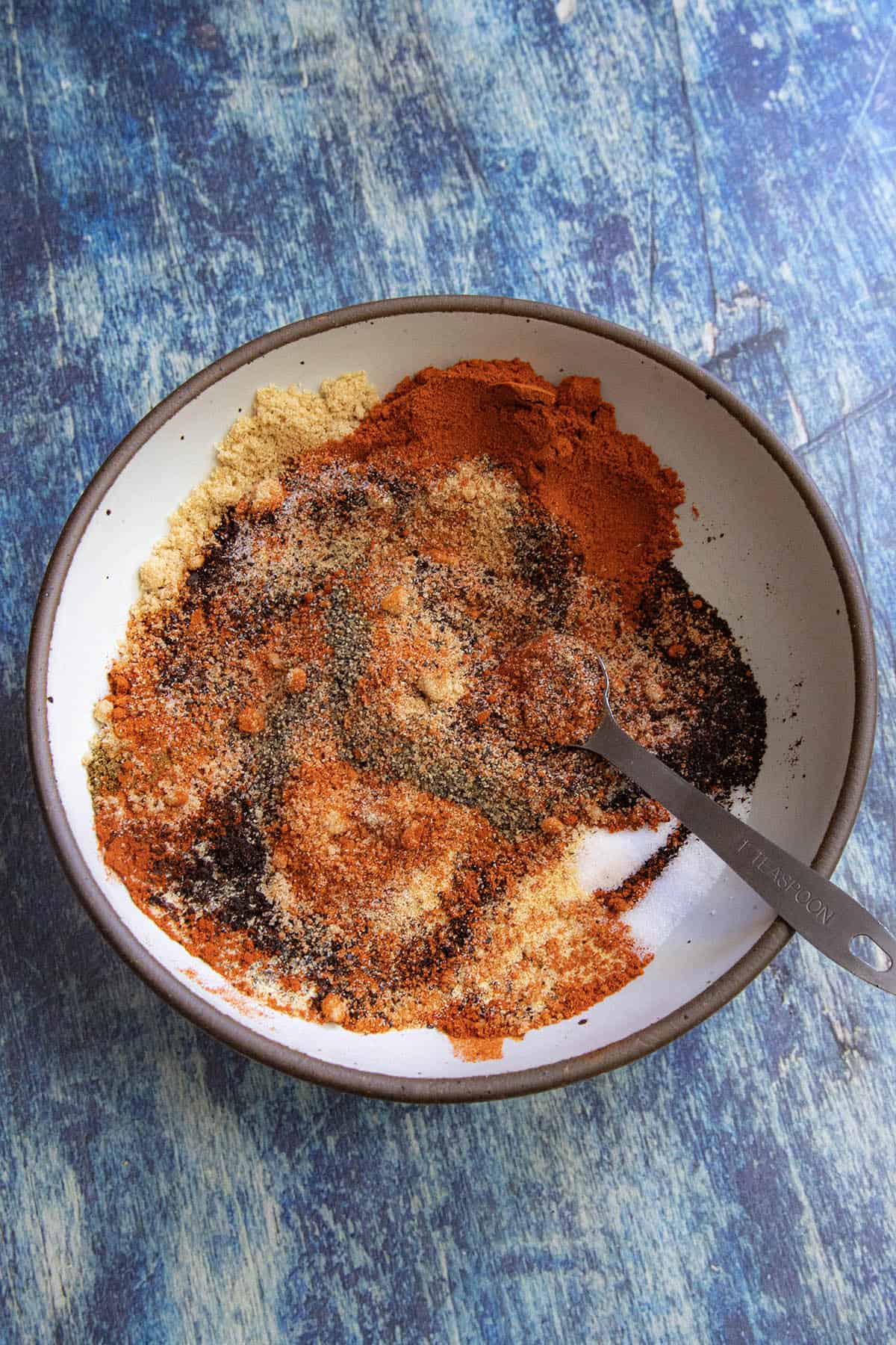 A bowl filled with various colorful ground spices and seasonings, partially mixed to create a spicy rib rub, with a metal measuring spoon resting in the mixture on a blue textured surface.