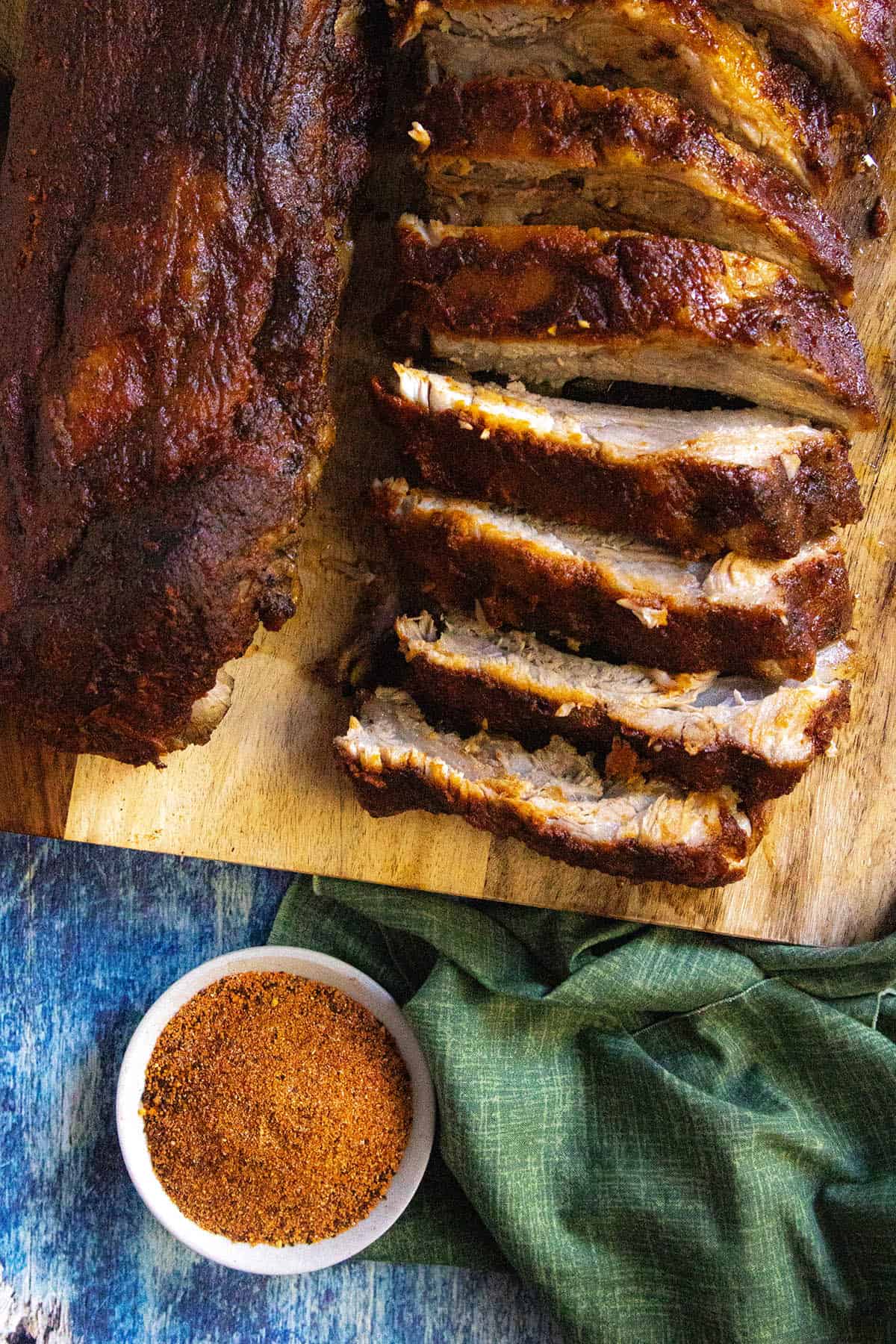 Overhead view of sliced barbecue pork ribs on a wooden cutting board, paired with a small bowl of dry rub for ribs seasoning and a green cloth on a blue surface.