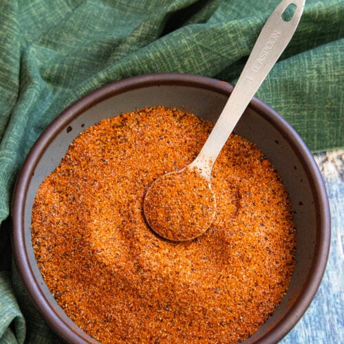 A bowl filled with reddish-brown homemade rib rub sits on a table, with a metal tablespoon resting on top. A green cloth is partially visible in the background.