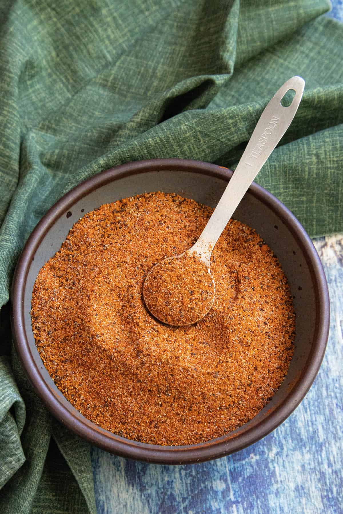 A bowl filled with reddish-brown homemade rib rub sits on a table, with a metal tablespoon resting on top. A green cloth is partially visible in the background.