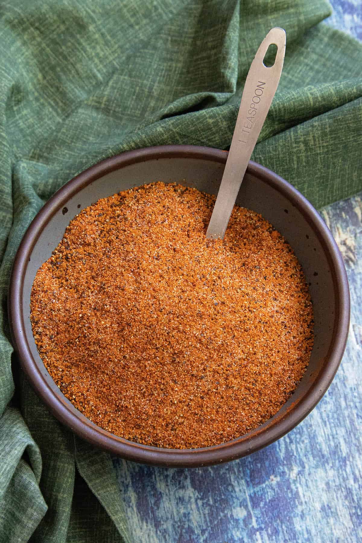 A brown bowl filled with orange-red spice mix, perfect for a rib rub recipe, with a metal measuring spoon labeled 1 TSP SPOON resting inside. The bowl sits on a green cloth over a blue textured surface.