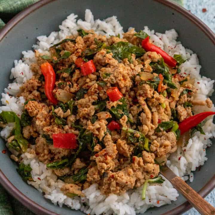 A bowl of white rice topped with Thai Basil Chicken, leafy greens, red Thai peppers, and spices, with a fork on the side. A green cloth is partially visible in the background.