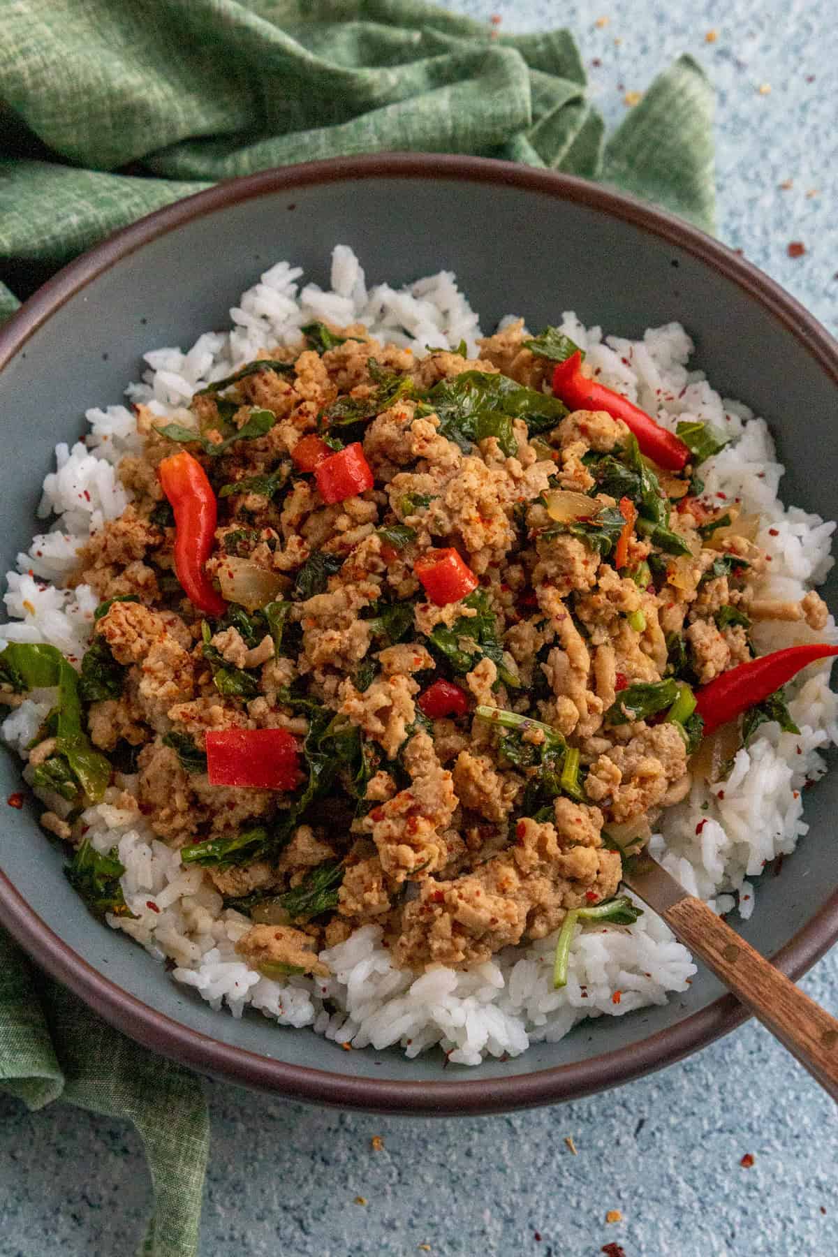 A bowl of white rice topped with Thai Basil Chicken, leafy greens, red Thai peppers, and spices, with a fork on the side. A green cloth is partially visible in the background.