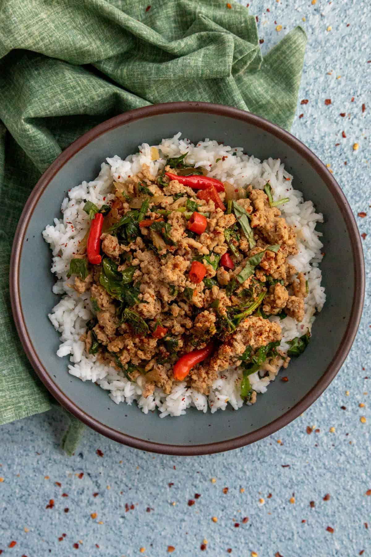 A bowl of white rice topped with Thai Basil Chicken, Thai pepper slices, and leafy greens, placed on a light blue surface with a green cloth napkin nearby.
