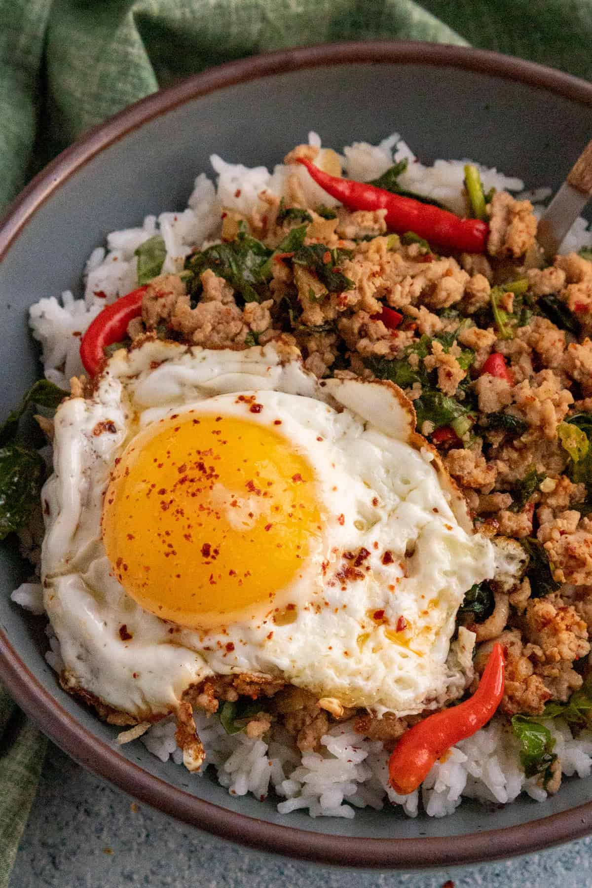 A bowl of white rice topped with Thai Basil Chicken, fresh herbs, red chili peppers, and a sunny-side-up fried egg sprinkled with chili flakes.