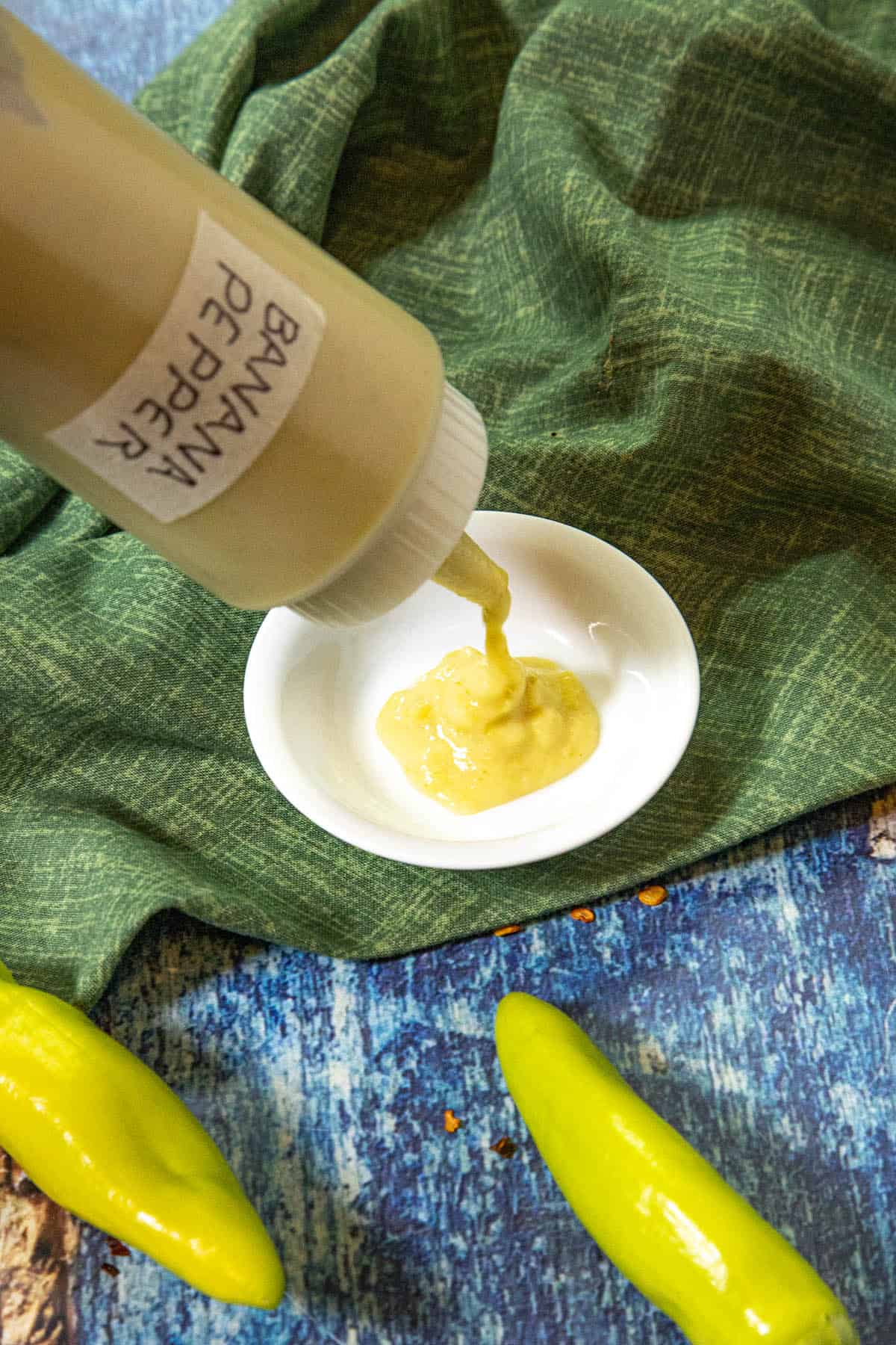 A bottle labeled Banana Pepper Hot Sauce pours vibrant yellow sauce into a small white bowl, with fresh banana peppers and a green cloth in the background on a blue surface.