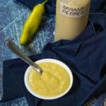 A small white bowl of creamy yellow Banana Pepper Hot Sauce with a spoon, next to a labeled bottle and a whole banana pepper, set on a dark blue cloth over a blue textured surface.