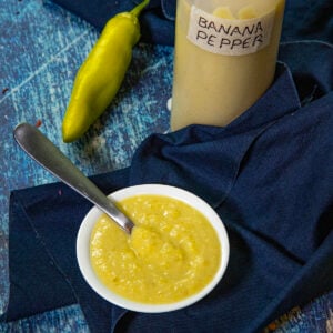 A small white bowl of creamy yellow Banana Pepper Hot Sauce with a spoon, next to a labeled bottle and a whole banana pepper, set on a dark blue cloth over a blue textured surface.