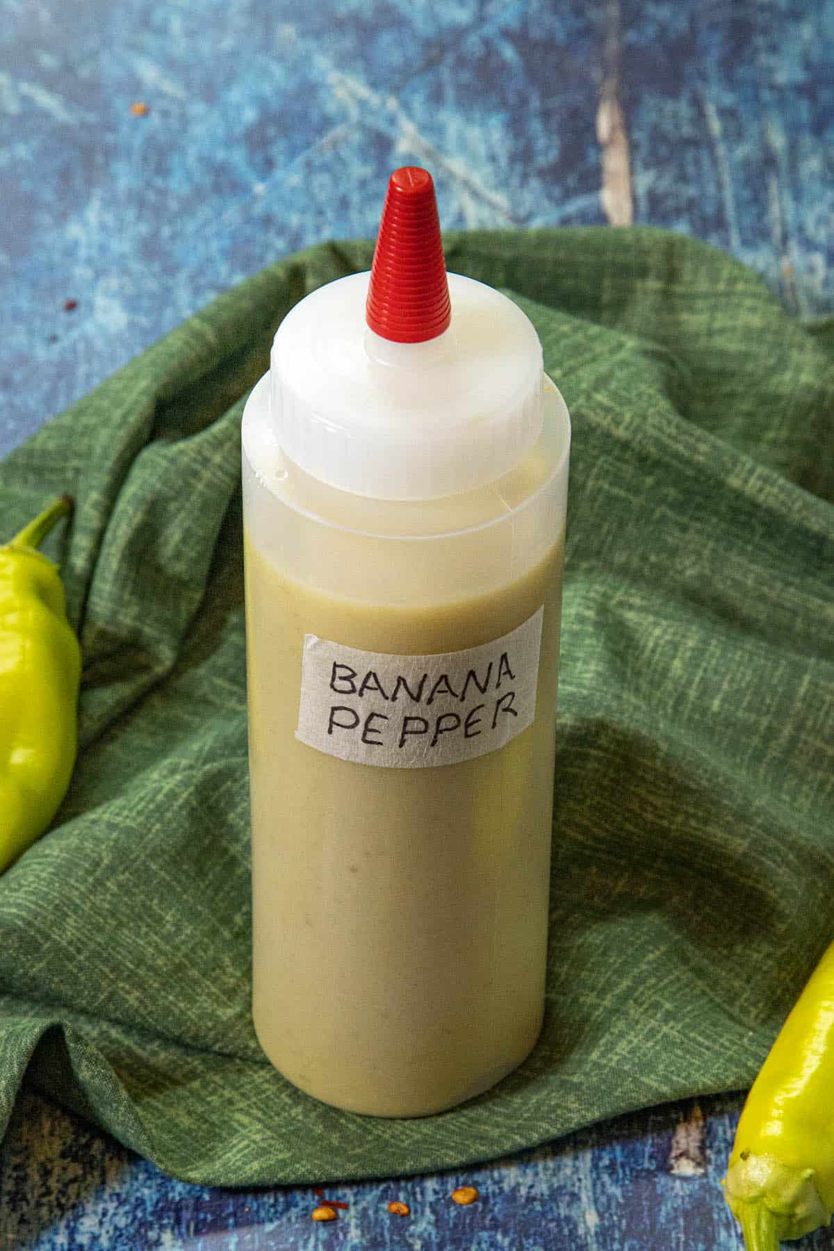A squeeze bottle with a red tip labeled Banana Pepper Hot Sauce sits on a green cloth, with yellow banana peppers nearby, against a blue textured background.