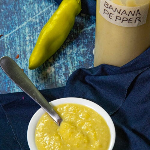 Small white bowl of creamy yellow banana pepper sauce with a spoon, next to a yellow banana pepper and a squeeze bottle labeled BANANA PEPPER HOT SAUCE, all set on a blue cloth and wooden surface.