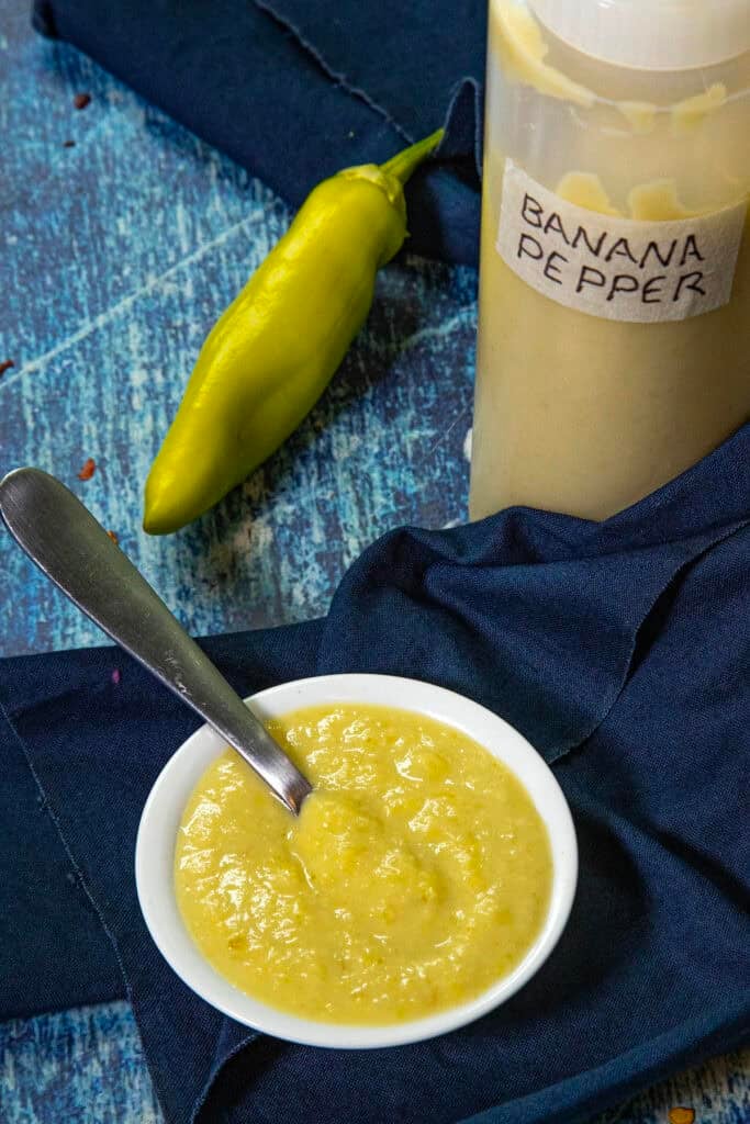 Small white bowl of creamy yellow banana pepper sauce with a spoon, next to a yellow banana pepper and a squeeze bottle labeled BANANA PEPPER HOT SAUCE, all set on a blue cloth and wooden surface.