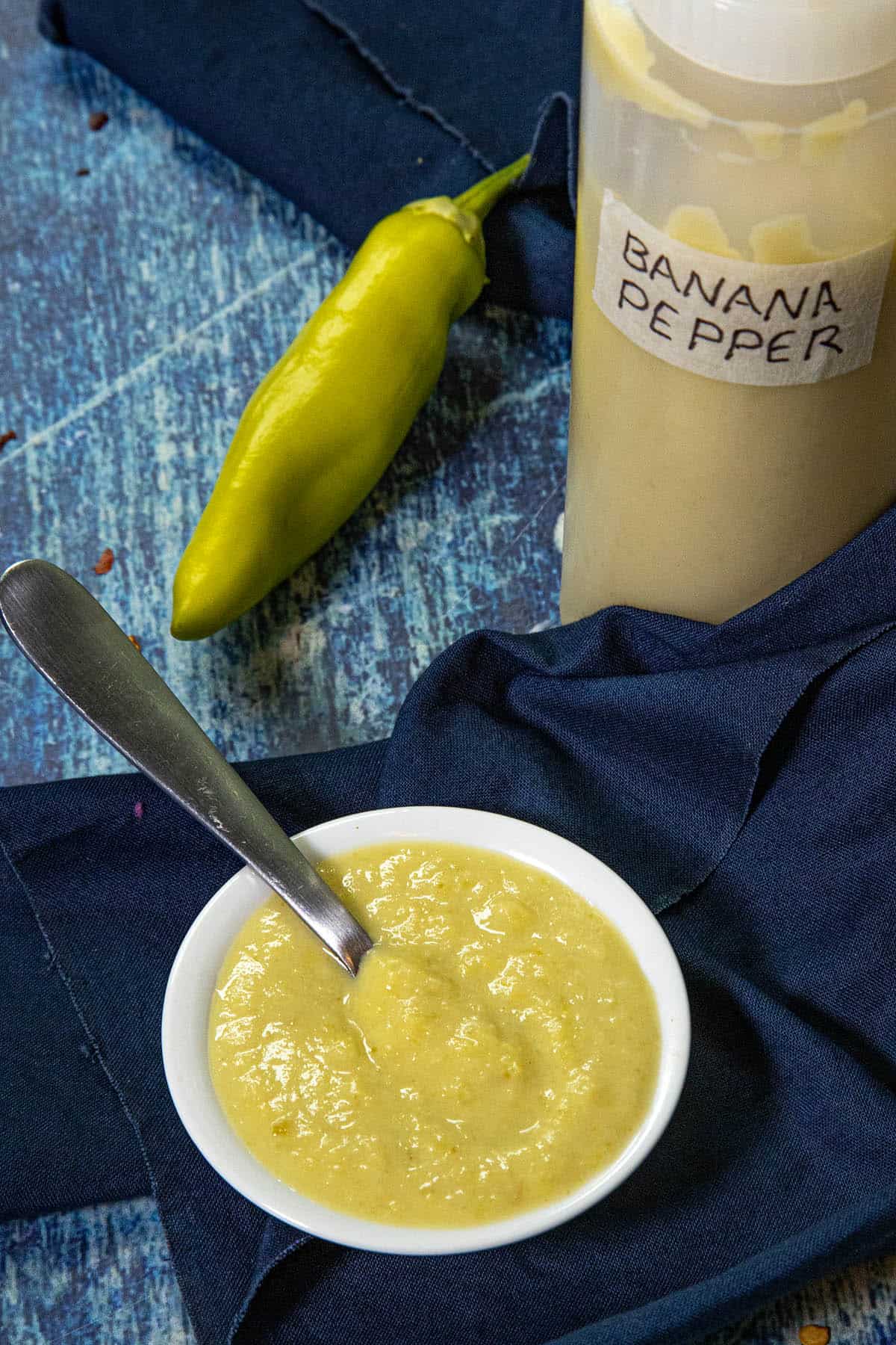 Small white bowl of creamy yellow banana pepper sauce with a spoon, next to a yellow banana pepper and a squeeze bottle labeled BANANA PEPPER HOT SAUCE, all set on a blue cloth and wooden surface.