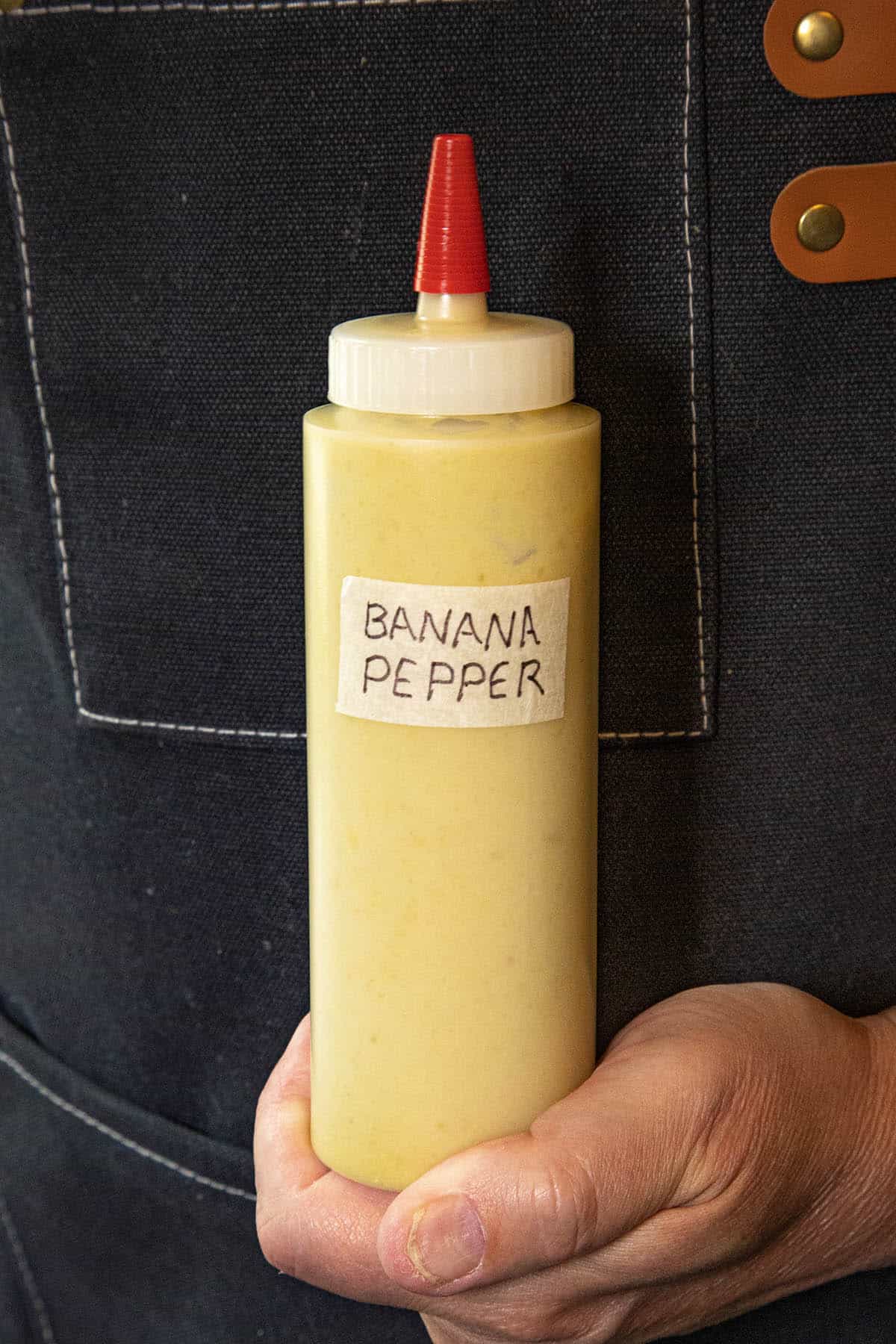 A hand holds a squeeze bottle with yellow sauce, labeled BANANA PEPPER HOT SAUCE, in front of a dark apron with brown leather accents.