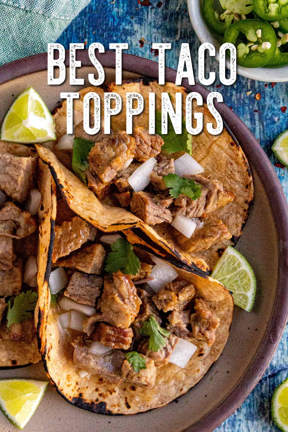 Three corn tortillas filled with chopped grilled meat, diced onions, and cilantro, served with lime wedges on a plate. Sliced jalapeños are visible in the background, highlighting some of the Best Taco Toppings. Text reads “Best Taco Toppings.”.