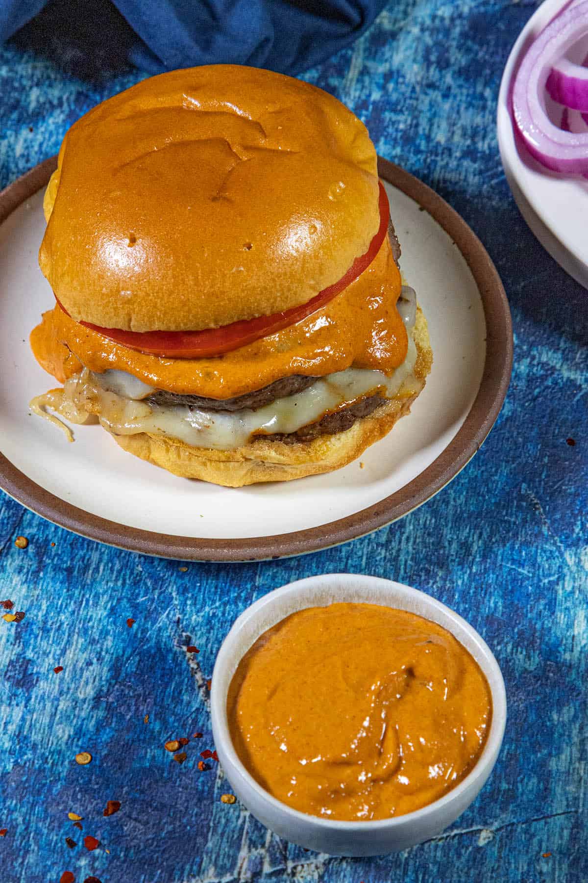A cheeseburger with melted cheese and a thick creamy burger sauce sits on a plate. Next to it is a small bowl filled with more of the same creamy burger sauce. Red onion slices are partially visible on another plate.