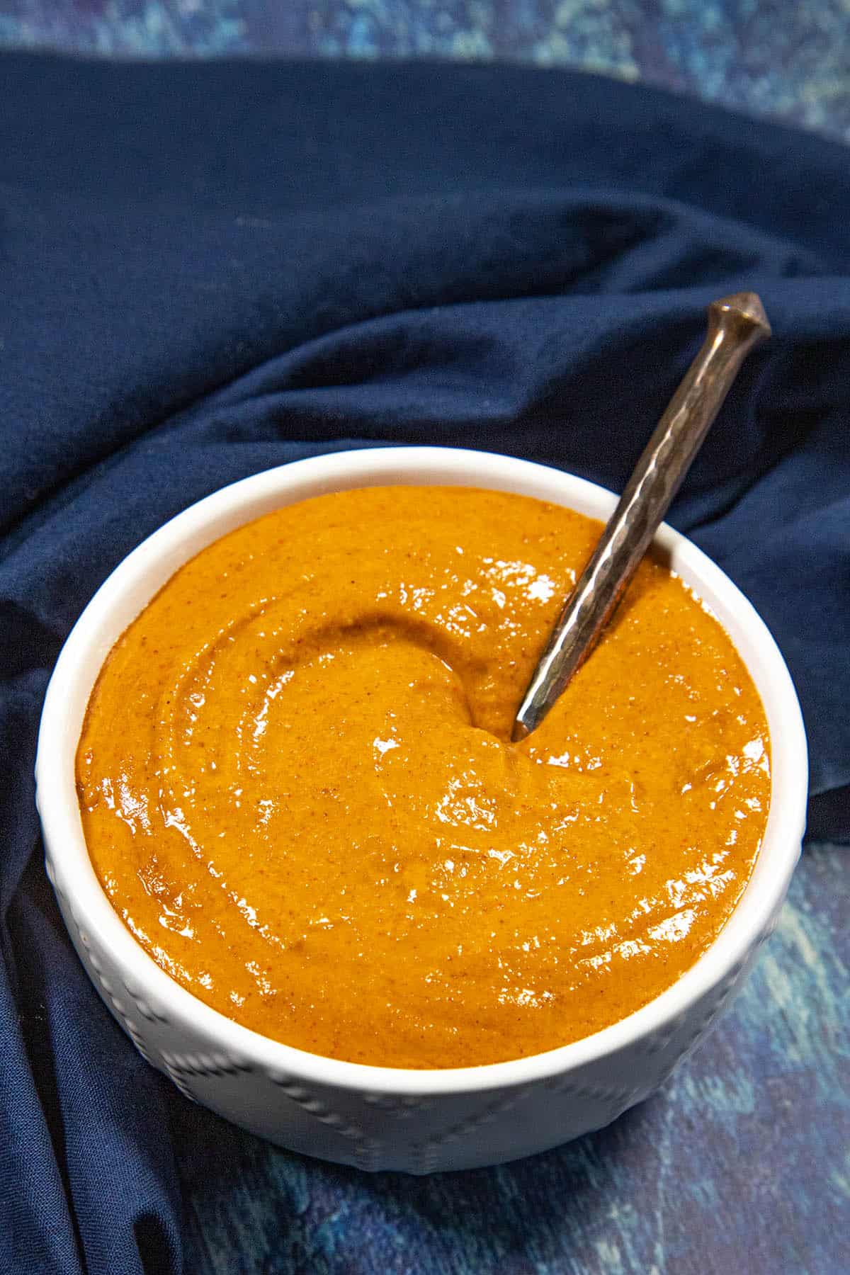 A white bowl filled with creamy burger sauce sits on a dark blue cloth, with a metal spoon resting inside the bowl.