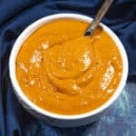 A white bowl filled with thick, reddish-orange Easy Creamy and Spicy Burger Sauce, with a spoon partially submerged, sitting on a dark blue cloth.