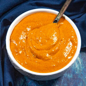 A white bowl filled with thick, reddish-orange Easy Creamy and Spicy Burger Sauce, with a spoon partially submerged, sitting on a dark blue cloth.