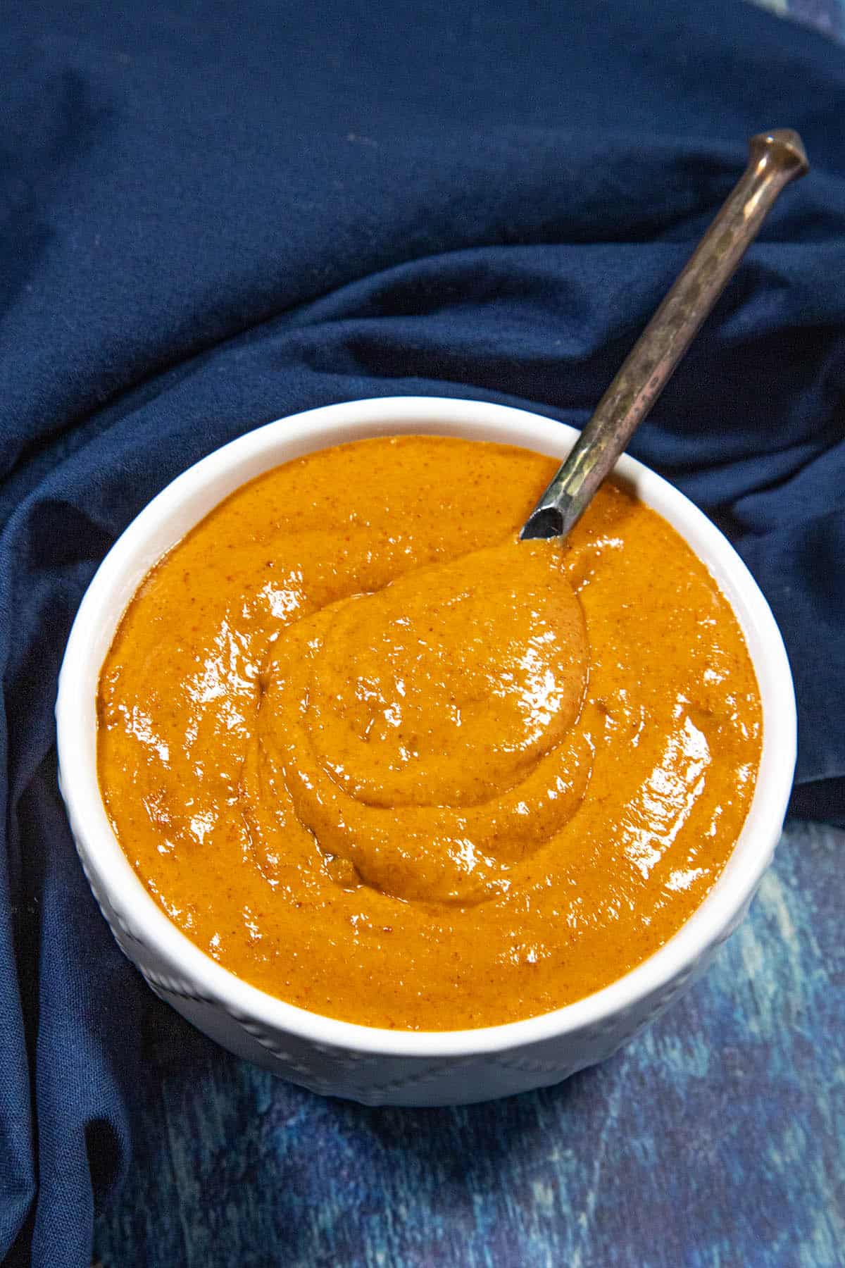 A white bowl filled with thick and creamy Spicy Burger Sauce, with a spoon resting inside. The bowl sits on a blue cloth on a blue textured surface.