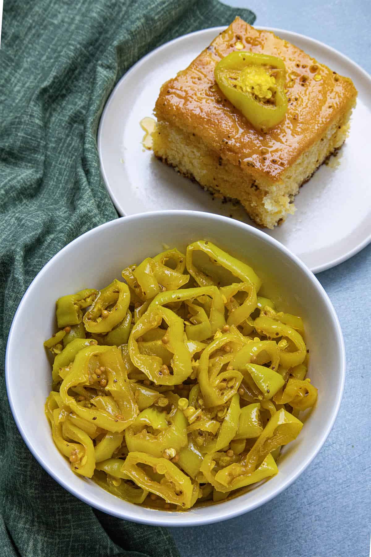A bowl of sliced, candied banana peppers sits next to a plate with a square slice of cornbread topped with a pepper slice, on a green cloth background.