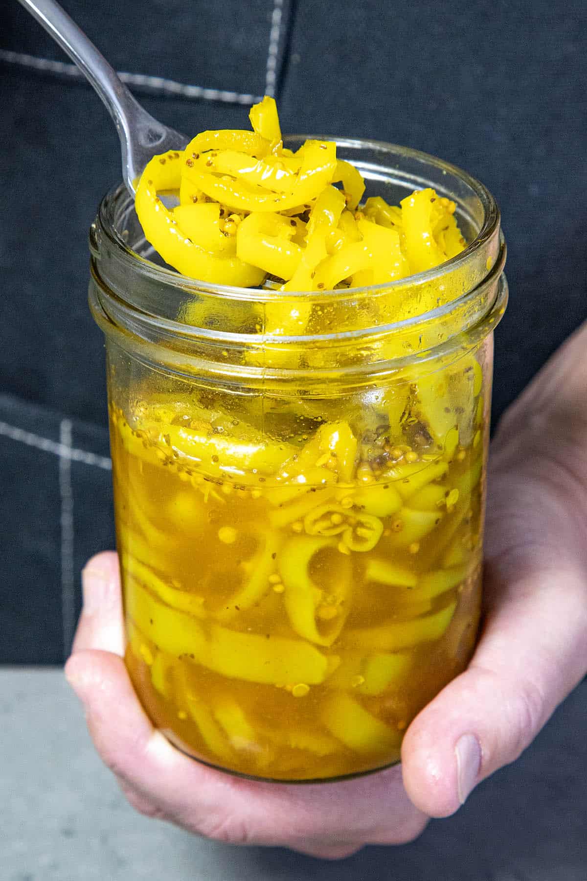 A hand holds a glass jar filled with yellow Candied Banana Peppers in brine. A spoon is scooping some peppers from the jar. The background is blurred and dark.