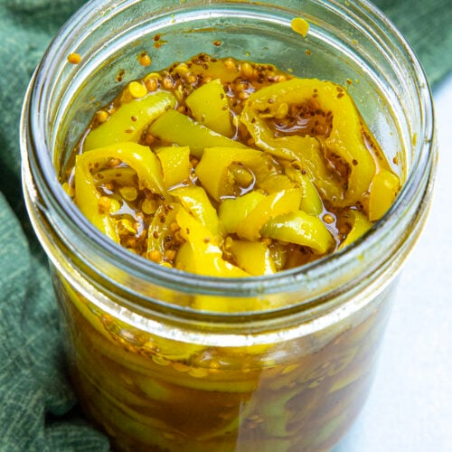 An open glass jar filled with yellow candied banana peppers and mustard seeds, sitting on a green cloth.