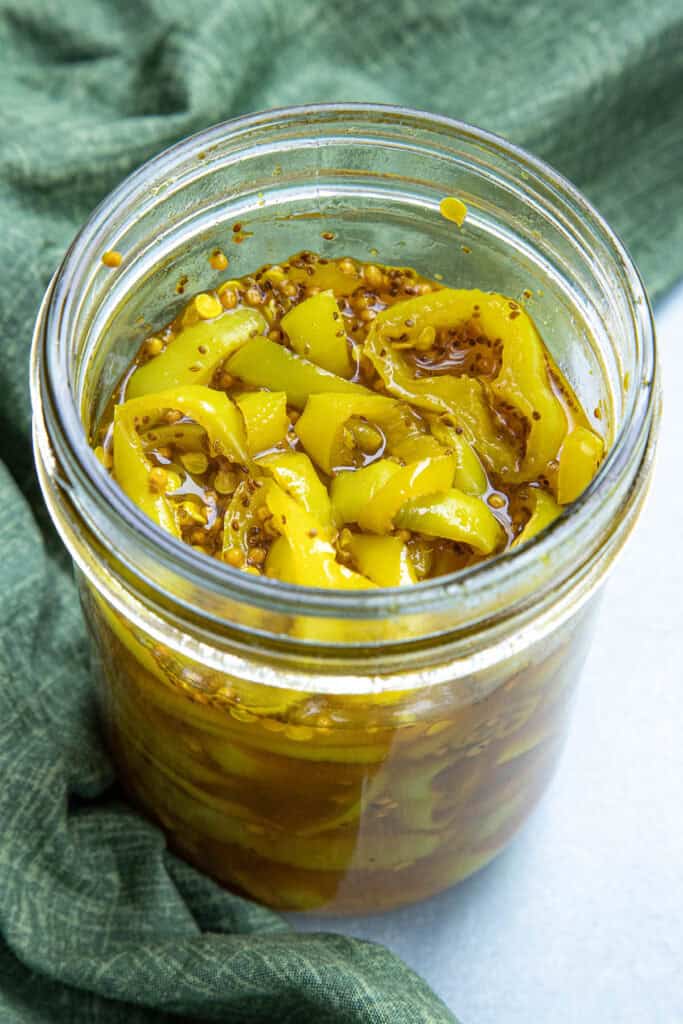 An open glass jar filled with yellow candied banana peppers and mustard seeds, sitting on a green cloth.