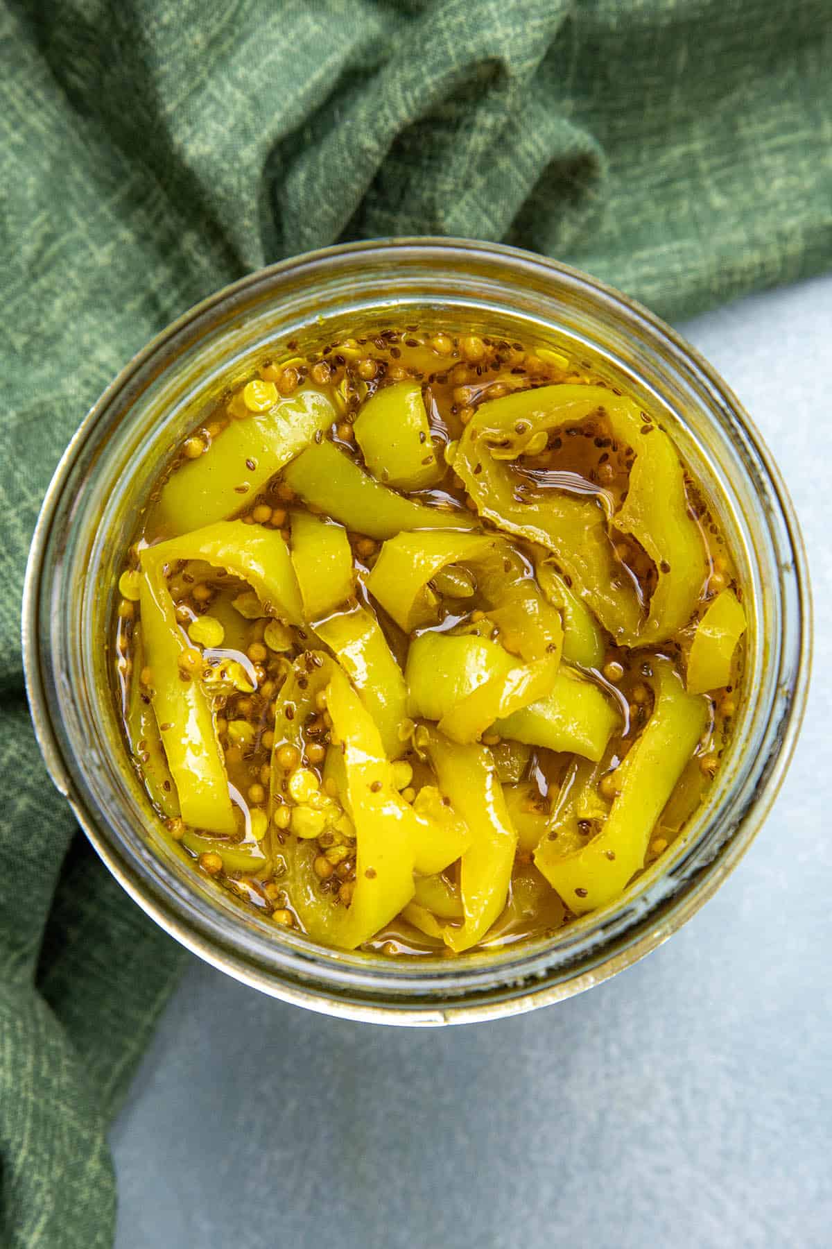 Overhead view of a glass jar filled with yellow pickled candied banana peppers in brine, with mustard seeds visible. The jar is placed on a green cloth, and the background is a light, neutral color.