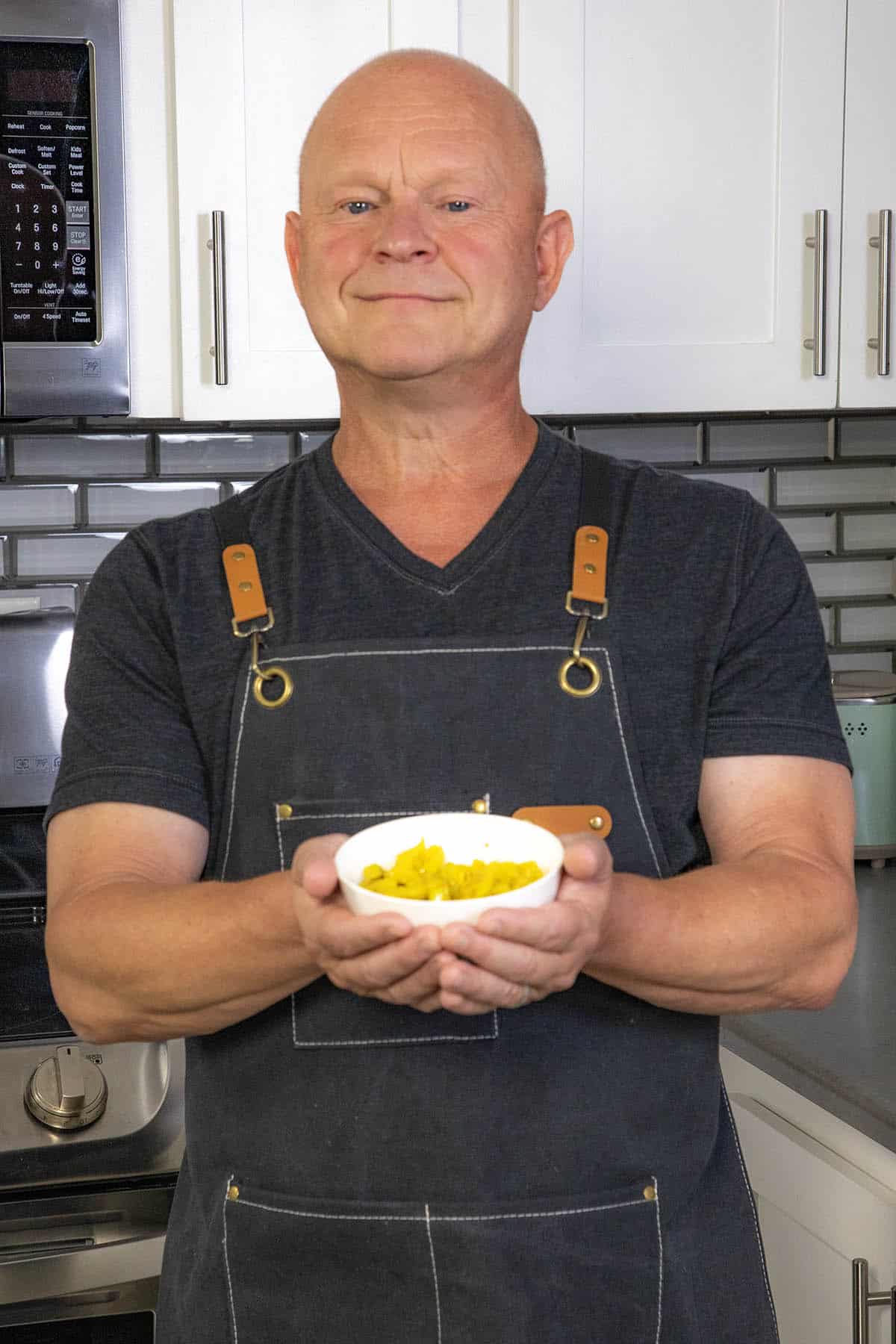 Mike from Chili Pepper Madness stands in a kitchen, holding a white bowl filled with candied banana peppers.