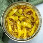 A top-down view of a jar filled with candied banana pepper rings in brine, with visible mustard seeds, on a green cloth background.