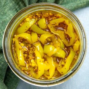 A top-down view of a jar filled with candied banana pepper rings in brine, with visible mustard seeds, on a green cloth background.