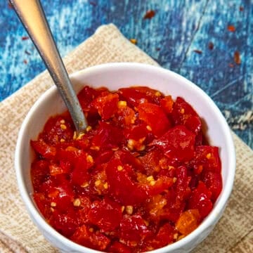 A white bowl of chopped red chili peppers with seeds, resembling a fresh Cherry Pepper Relish, sits with a spoon inside on folded beige napkins against a textured blue background.