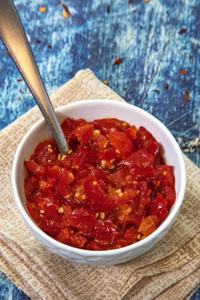 A white bowl of chopped red chili peppers with seeds, resembling a fresh Cherry Pepper Relish, sits with a spoon inside on folded beige napkins against a textured blue background.