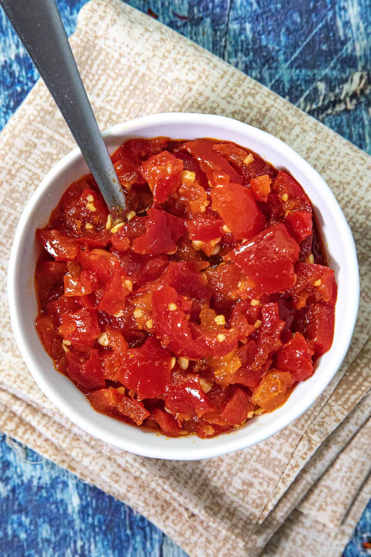 A white bowl filled with chopped red chili peppers and Cherry Pepper Relish sits on a folded beige napkin. A spoon is in the bowl, and the background features a textured blue surface.