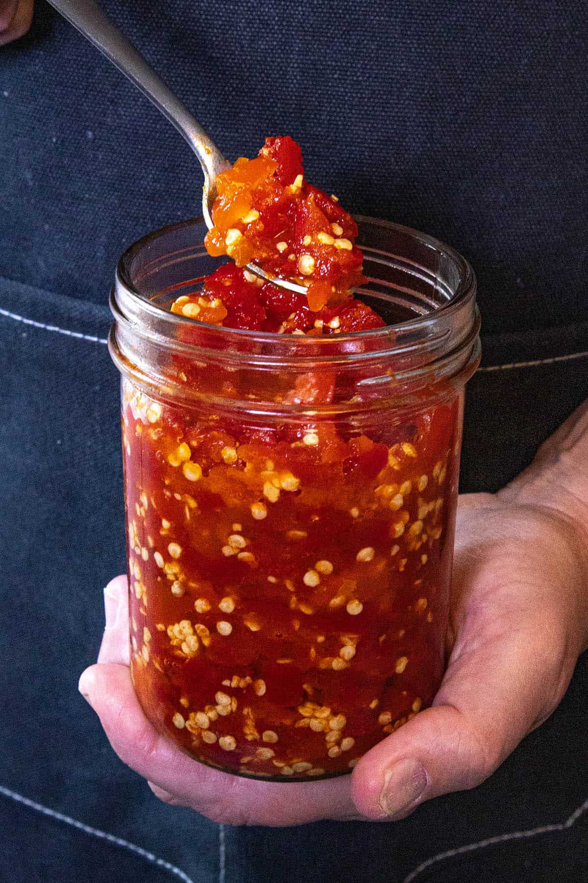 A person holding a jar filled with chunky red chili sauce, scooping some out with a spoon. The sauce, reminiscent of a vibrant Cherry Pepper Relish, contains visible chili seeds and pieces of red pepper.