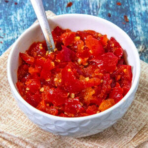 A white bowl filled with vibrant Cherry Pepper Relish, with a spoon inside, sits on a beige napkin and a blue wooden surface.