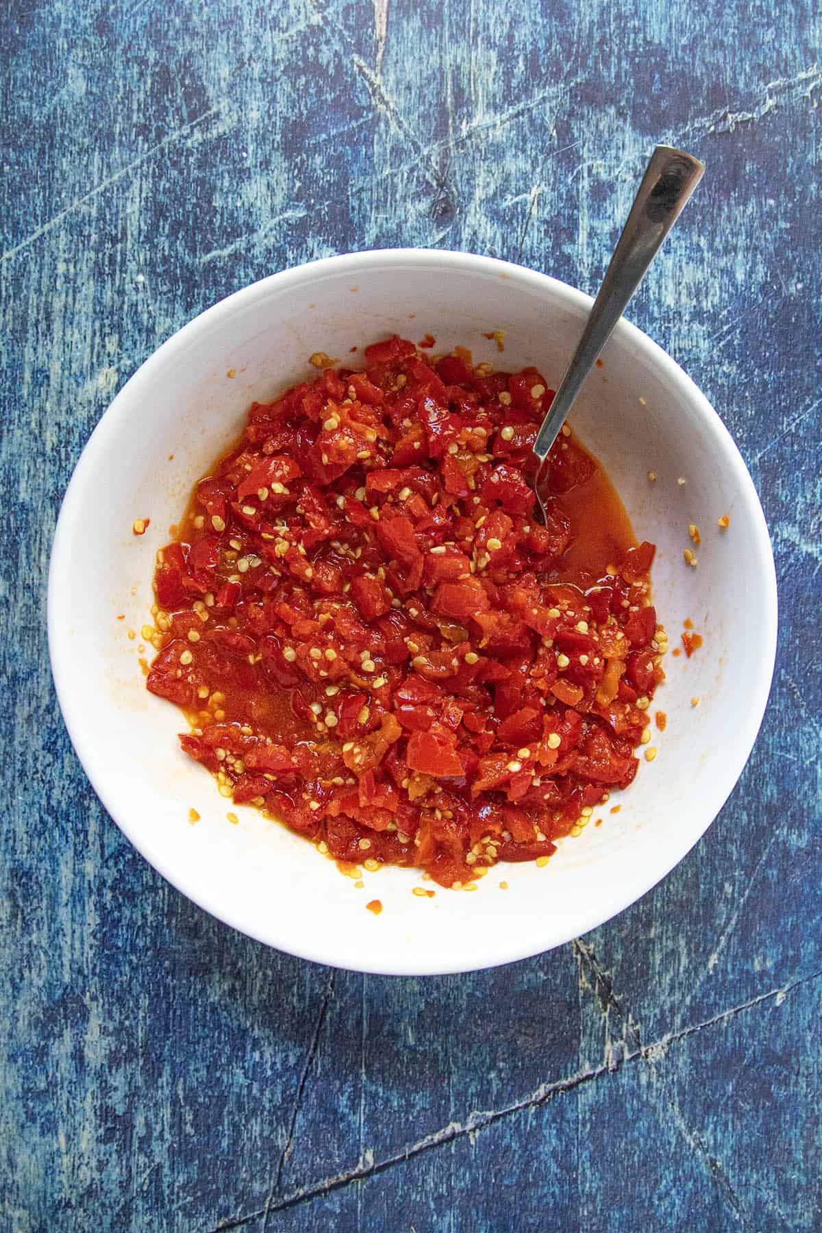 A white bowl filled with finely chopped red chili peppers and seeds, resembling a vibrant Cherry Pepper Relish, with a spoon inside, sits on a blue textured surface.