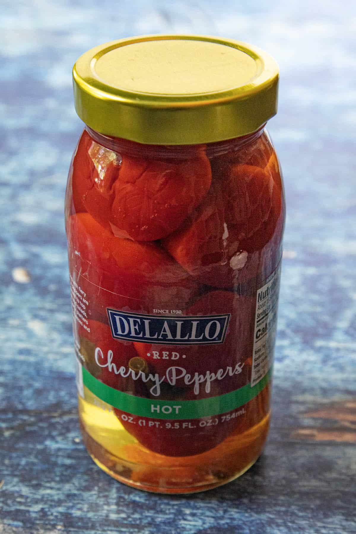 A jar of DeLallo Red Cherry Peppers labeled Hot sits on a blue textured surface. The jar, perfect for creating a zesty Cherry Pepper Relish, is filled with bright red peppers and has a yellow lid.