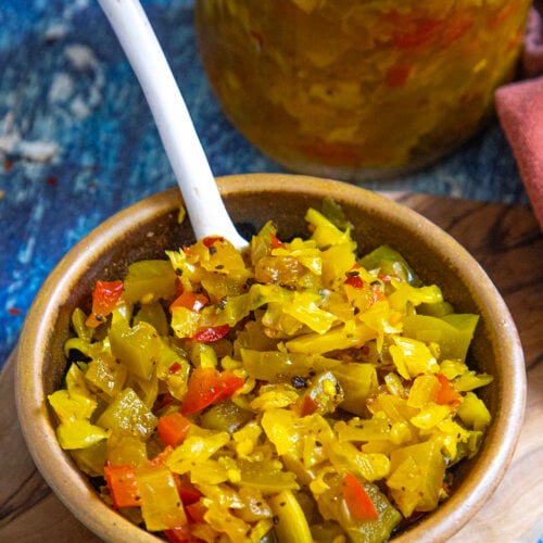A brown bowl filled with colorful chopped Chow Chow Relish, featuring yellow, green, and red pieces, sits on a wooden board with a spoon. A jar of the same relish is visible in the background on a blue surface.