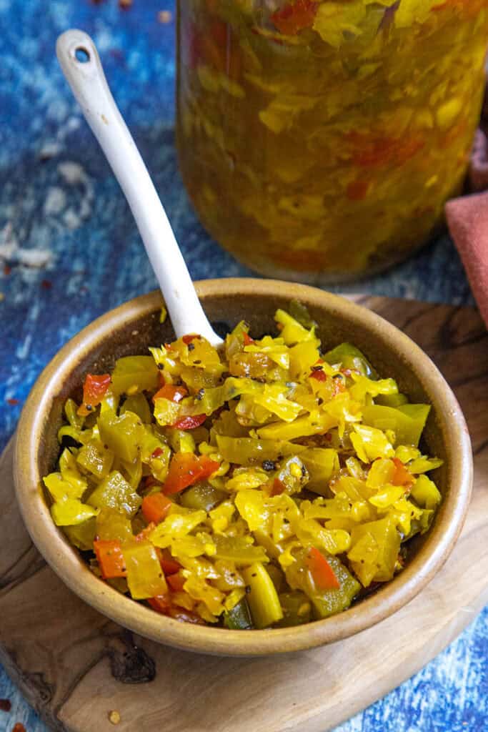 A brown bowl filled with colorful chopped Chow Chow Relish, featuring yellow, green, and red pieces, sits on a wooden board with a spoon. A jar of the same relish is visible in the background on a blue surface.