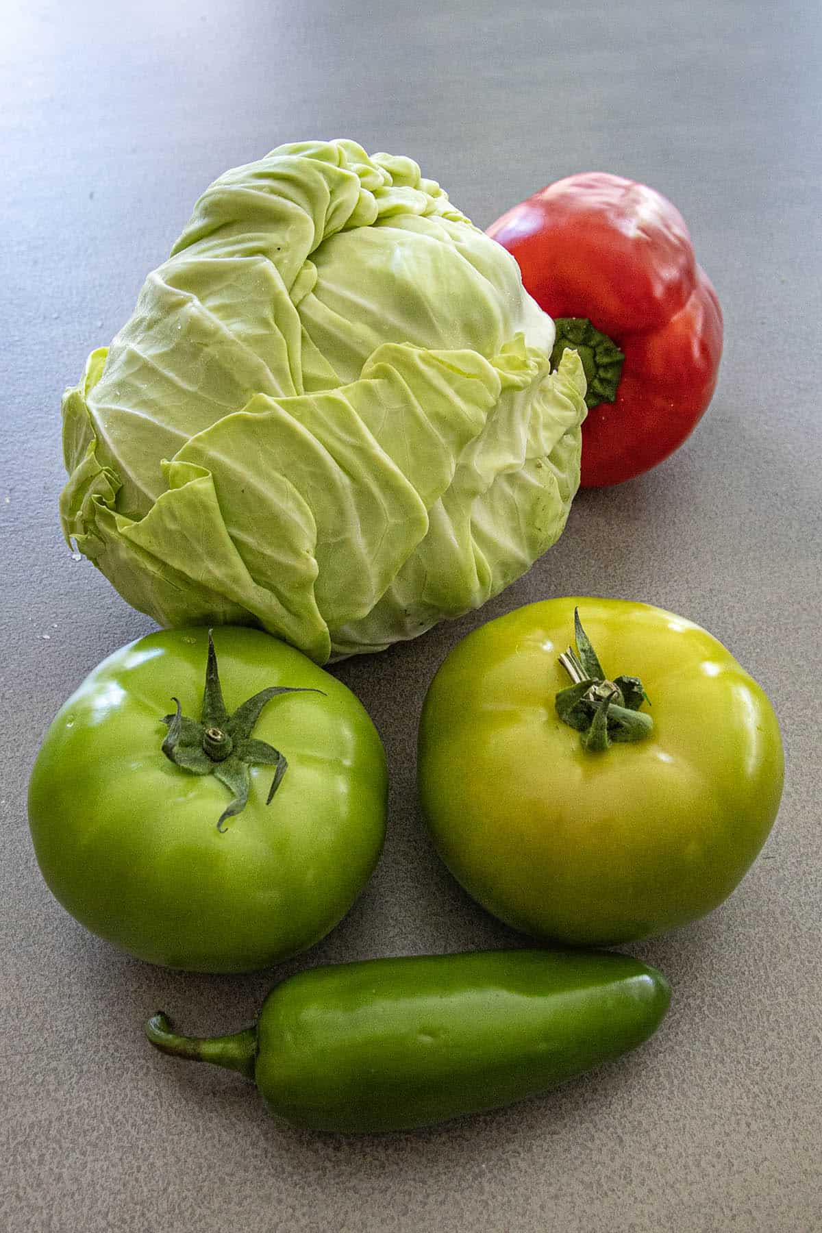 A head of cabbage, a red tomato, two green tomatoes, and a jalapeño pepper—classic Chow Chow Relish ingredients—are arranged on a gray surface.